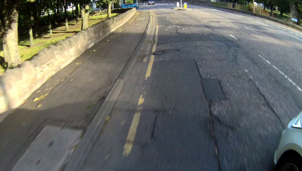 Alongside the traffic island. The front of a vehicle is visible to the right, between the cyclist and traffic island.