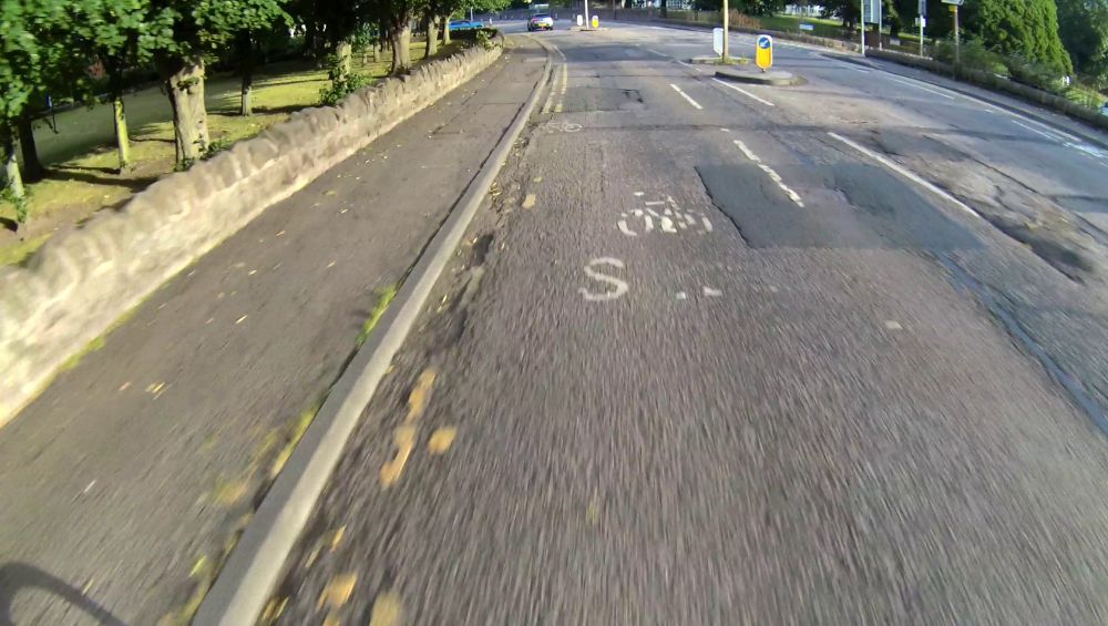 A clear road with a wide cycle lane and narrow general traffic lane, on the approach to a traffic island.