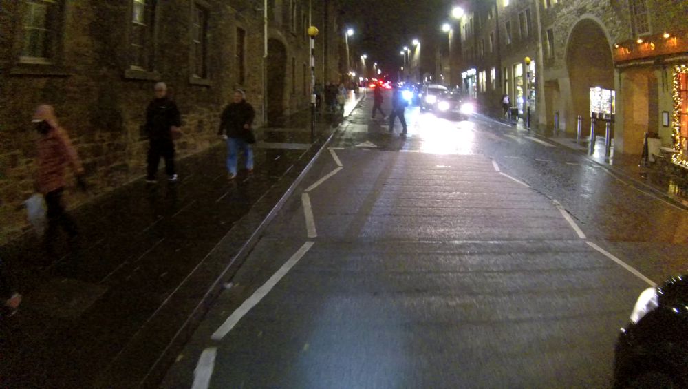 A cyclist approaching a zebra crossing in primary position (middle of the lane), around 8 metres away. There are pedestrians crossing. The front left wing of an overtaking taxi is visible in the lower right of the image.