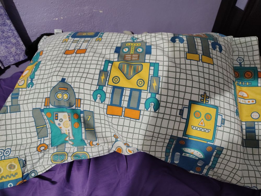 A pillow with a case depicting various toy robots over a grid background sitting on a bed with purple sheets