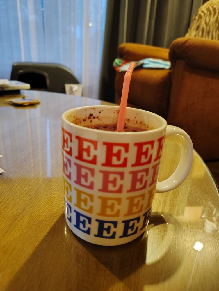 A mug of hot cocoa on a table, the mug is white and covered in letter Es of various colors.