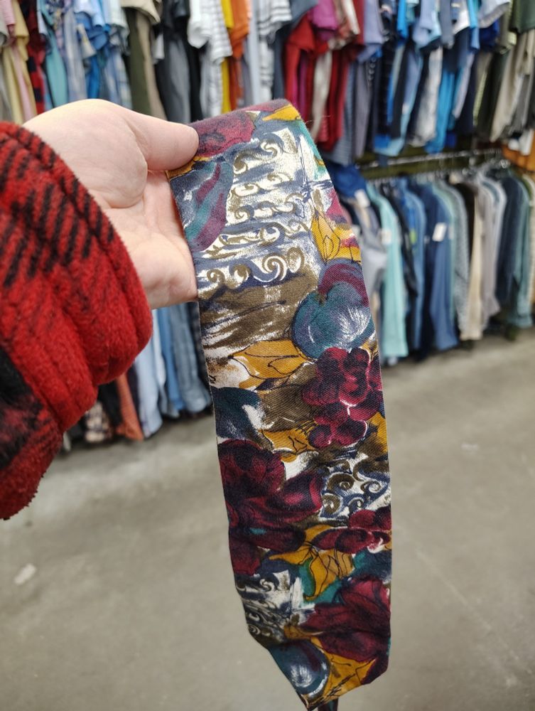 A tie with abstract, paint like patterns in gold, beige, blue, and magenta. It's being held aloft in front of a rack of other thrift store clothes. 