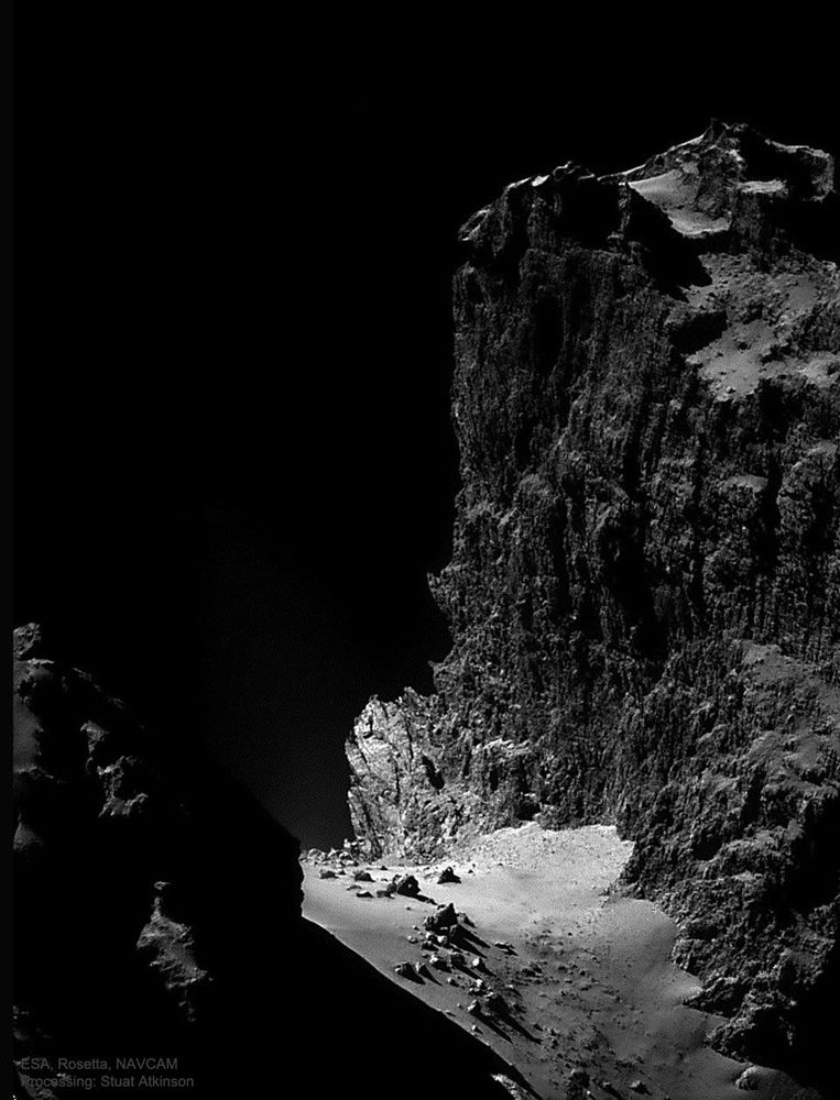 A black-and-white photo of a high, rocky cliff on a comet. Icy sand pools at the bottom of the cliff and is scattered with large boulders. The black of space provides the backdrop.