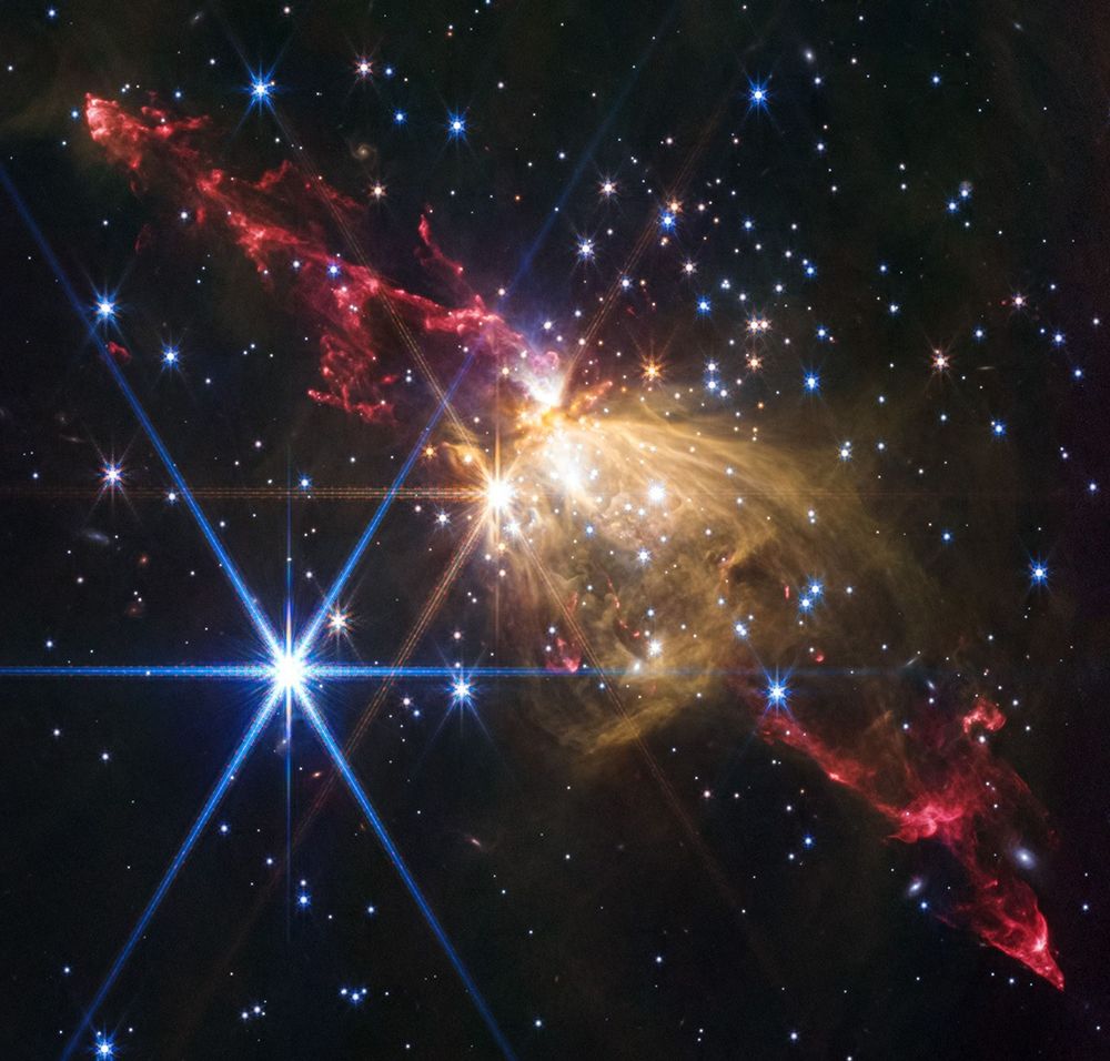 A red jet of gas stretches across the image from the upper left to the lower right. The center of the image contains a mass of gold gas and dust the blocks the jet from view. Two bright stars, one blue and one yellow, are in front of the jets and show the tell-tale six-way stellar spikes of JWST. Other smaller stars dot the background and foreground.