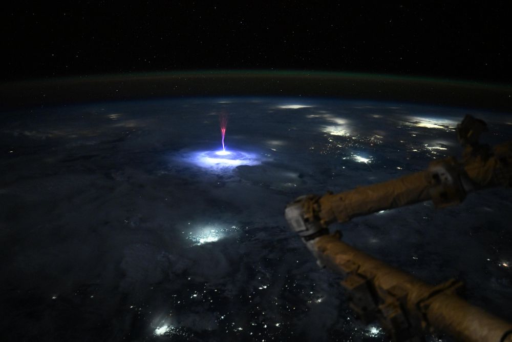 Taken from the ISS, a gigantic jet spears up from a thunderstorm cloud near the Texas-Mexico border. The spear is blue-white at the cloud, with a massive light spreading out in all directions. Near the top of the jet, the lightning turns to red and branches out. In the foreground is the Canadarm2. The city lights of Dallas, Austin, San Antonio, and Houston are to the northeast, and Torreón, Mexico, to the southwest.
