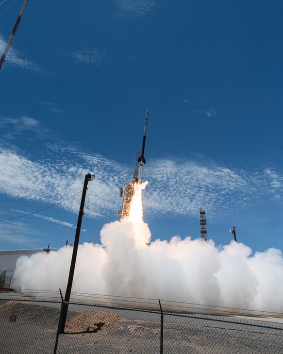 A sounding rocket launches into a nearly cloudless blue sky, a plume of smoke billowing out along the ground from below.