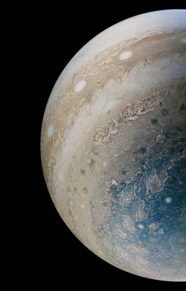 A view of one half of Jupiter, lit from the left, revealing alternating bands of white and tan full of swirling storms. The polar end of the planet is seen in shades of blue with more clouds swirling.