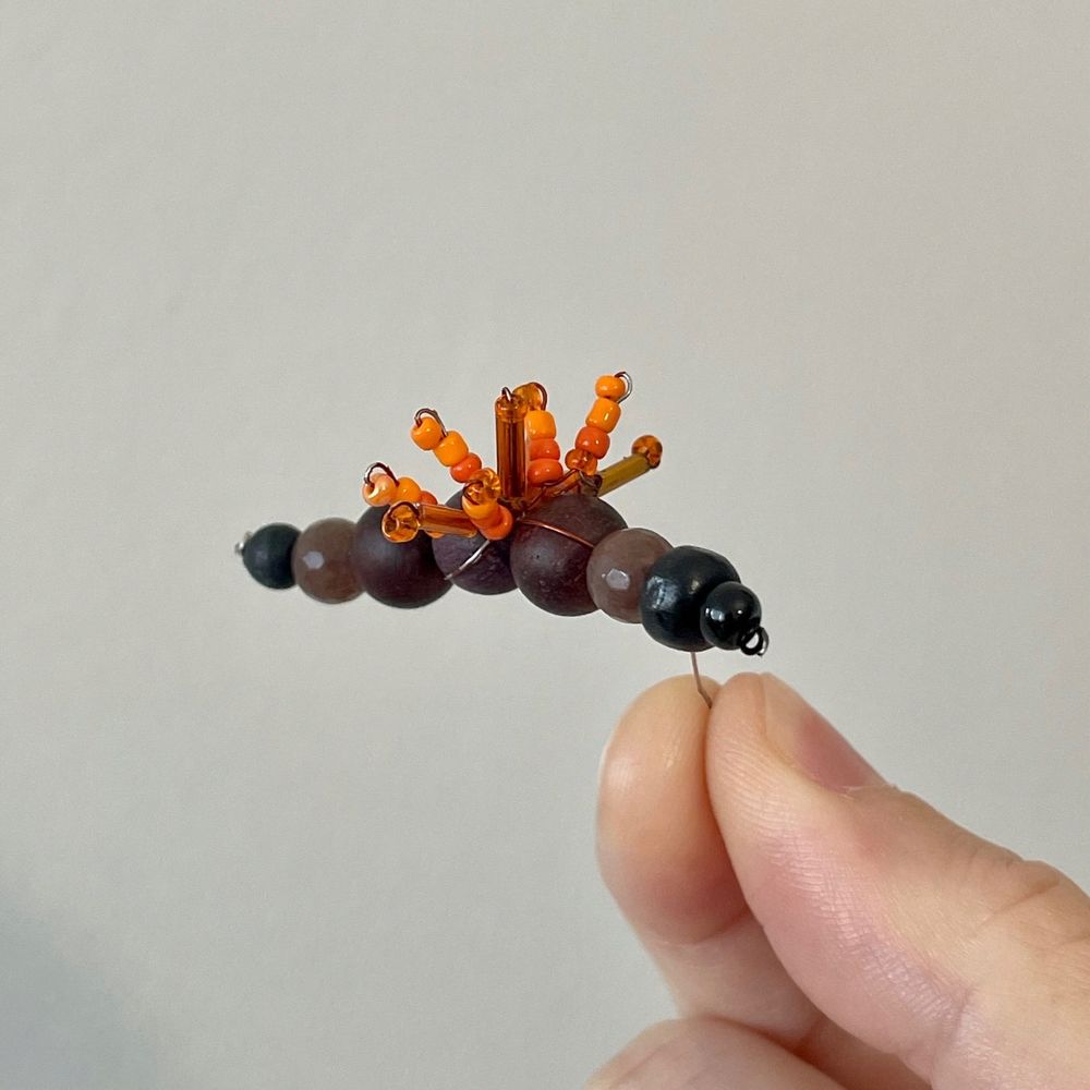 Holding a tiny work-in-progress sculpture of a woolly bear caterpillar; the bare body is made up of brown and black beads, and I just started adding orange beads around the center to represent the caterpillar's fuzzy bristles.