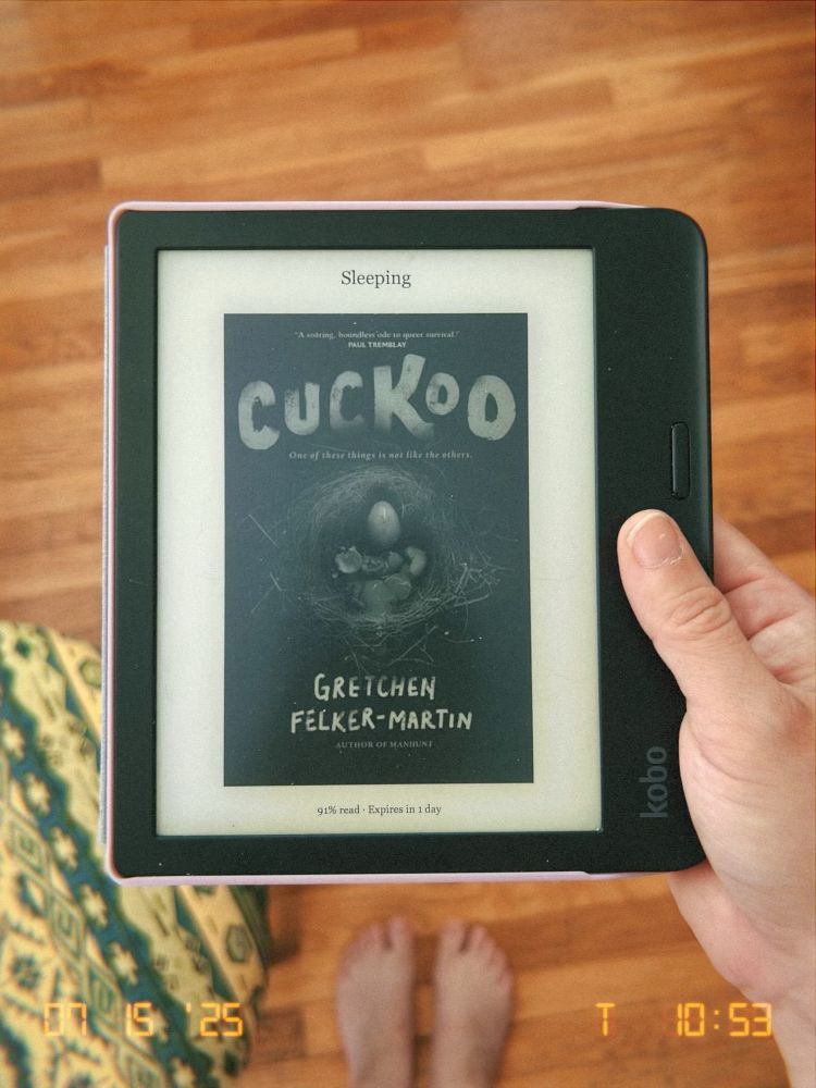 my hand holding an e-reader with the display showing the cover Cuckoo by Gretchen Felker-Martin. my feet on warm, hardwood floors are in the background out of focus next to a green and yellow geometric-patterned foot stool. 