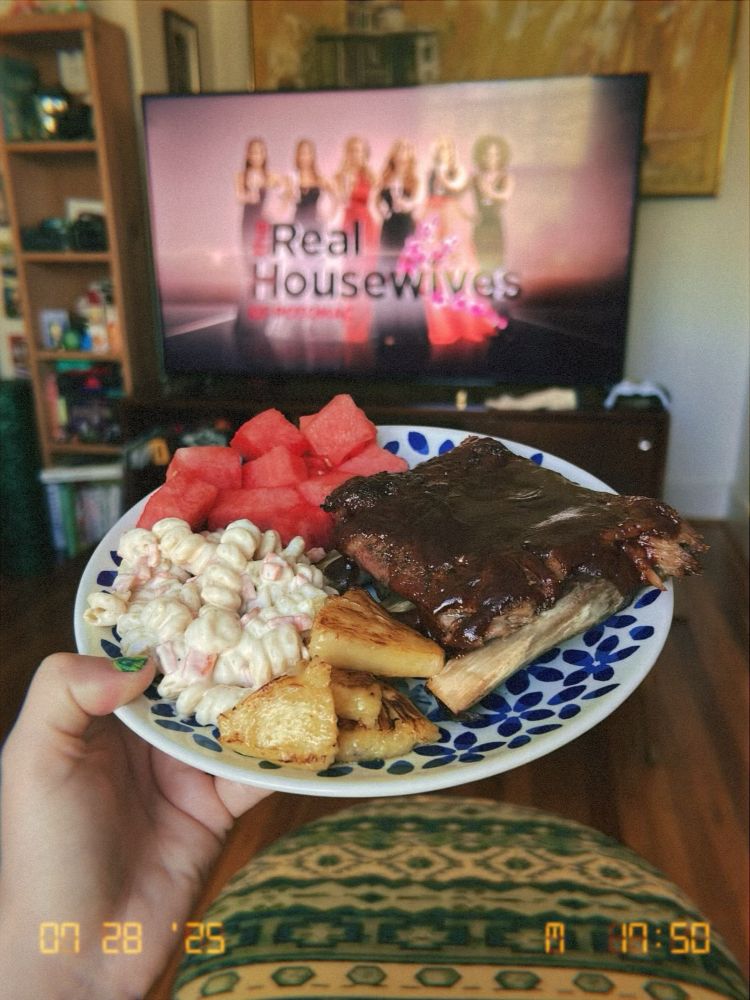 my hand holding a plate full of food (ribs slathered in dark bbq sauce, cubes of watermelon, mac salad, and grilled pineapple) with a tv in the background showing the intro to Real Housewives of Potomac. 