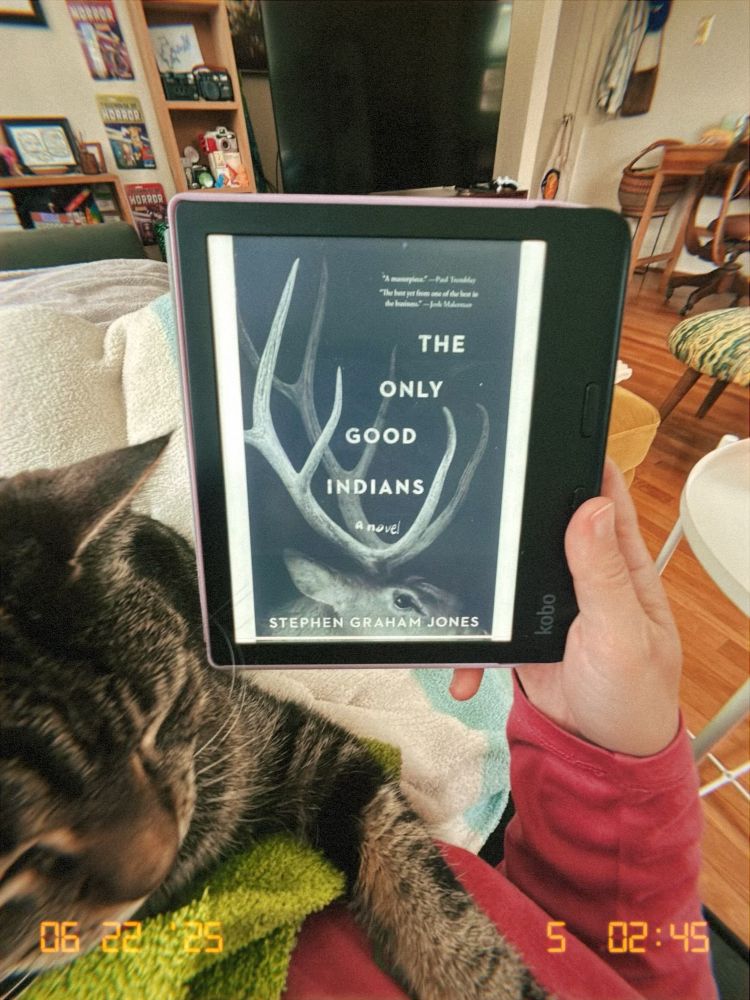 my hand holding an e-reader displaying the cover page for The Only Good Indians by Stephen Graham Jones. my tabby cat is laying on my chest in the foreground. 