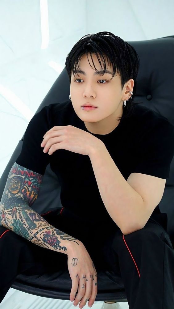 Jungkook sitting in a black leather chair posing for a photoshoot. He has a short sleeve black shirt on, tattoos exposed, and he looks very sexy.