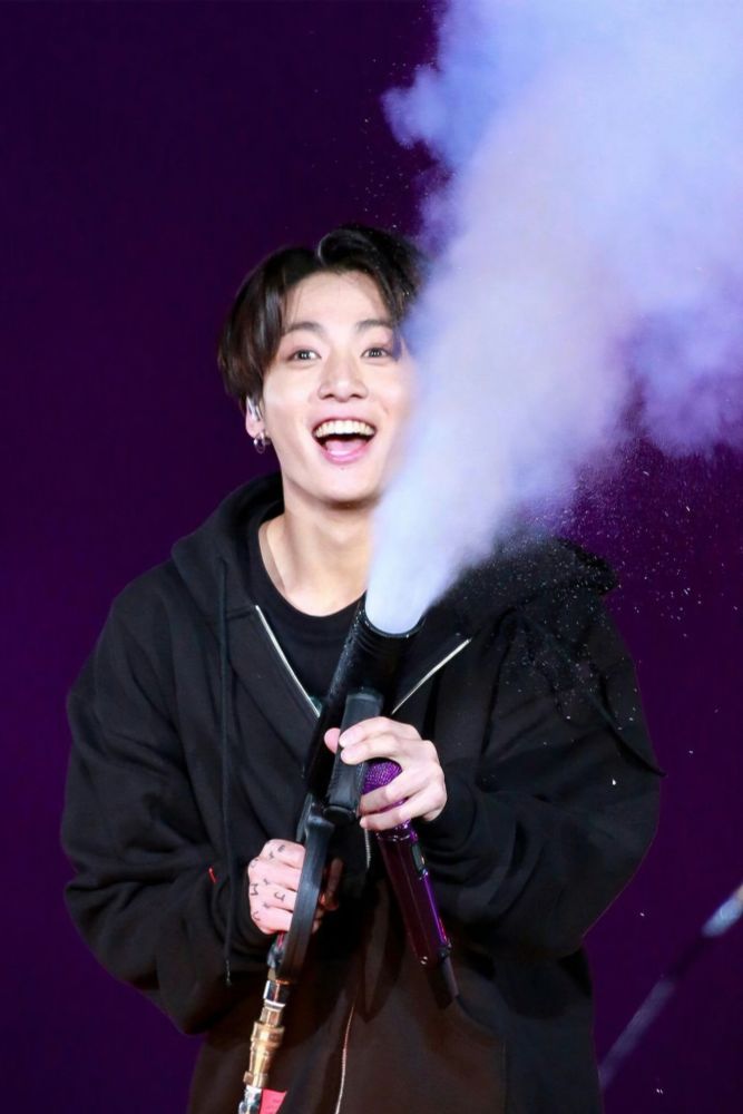 Jungkook on stage during a concert in a black hoodie spraying a mist gun.