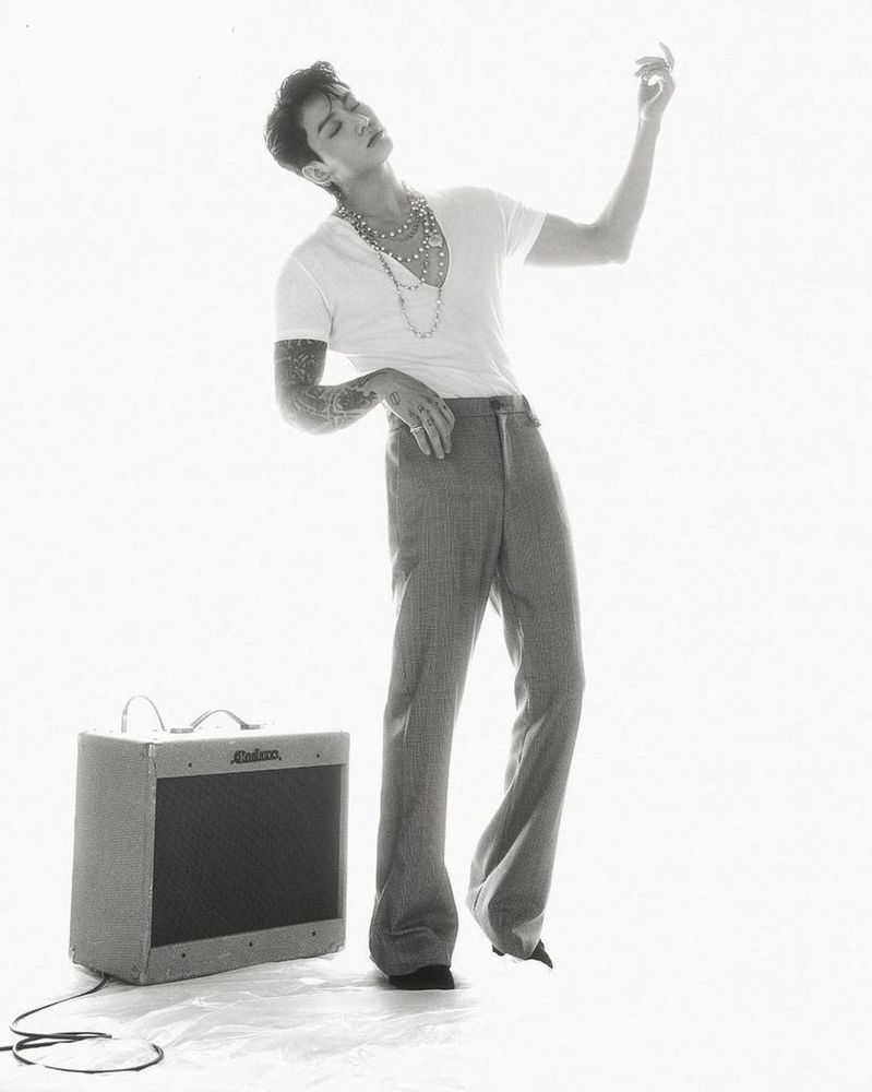 A black and white Calvin Klein photo of Jungkook in a white v neck T-shirt and bell bottom jeans strumming an air guitar.