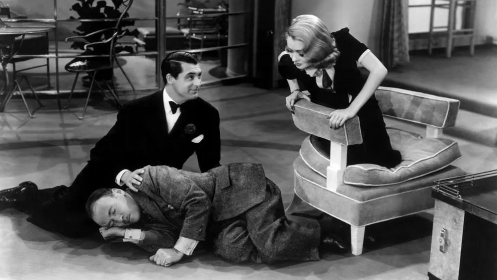 A still from Topper (1937). Cary Grant is dressed in a tuxedo and sitting on the floor with his legs out to the side. Roland Young wearing a grey suit and is asleep with his hand under his cheek and his head in Cary's lap. Constance Bennett is  wearing a black dress and kneeling on a chair looking down at the two men. 