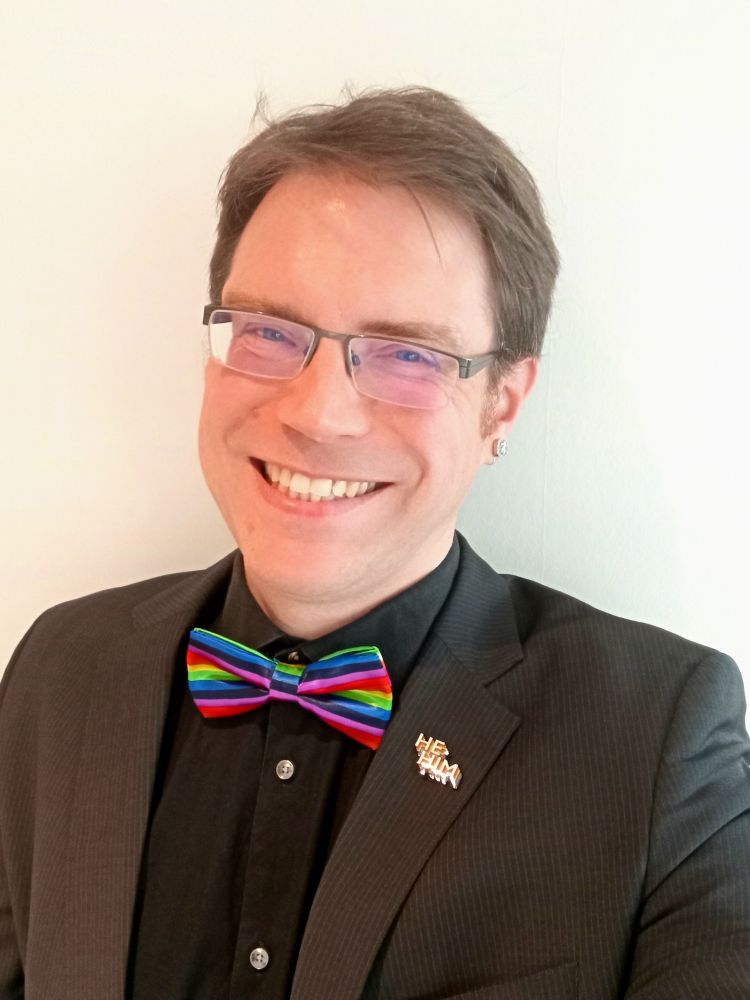 Self portrait of me, a middle aged white man. I'm wearing sparkly earrings, a black shirt, a rainbow striped bow tie and a black suit jacket with pinstripes.
There's a "he him" pin on the lapel of the suit jacket.