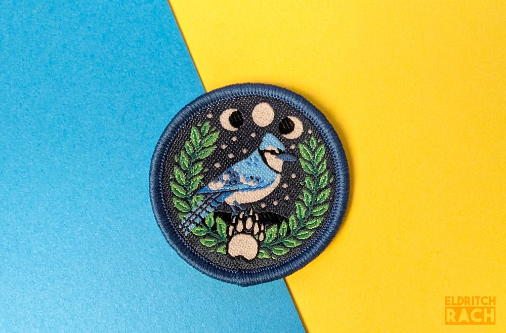 Woven patch - a blue jay sits on a laurel. There are 3 moons in the background and bear paw print in the foreground. Symbols of the druid from DND