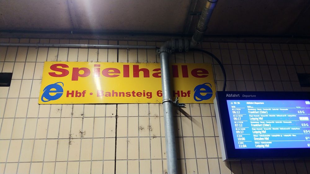 Photo of a tiled and partially roofed wall on the outside of Magdeburg train station. There's a large sign on the wall reading "Casino, main station, platform 6" with an old Internet Explorer logo (1997 - 2001) on both sides of the sign. The sign is partially obstructed by a drainpipe running downwards in front of it. On the right, a train departure screen is mounted to the wall.