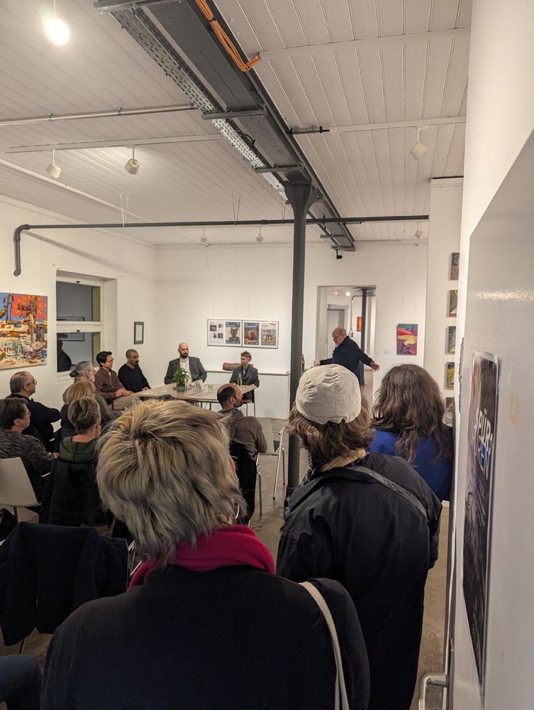 Eine Gruppe von Menschen sitzt in einem hellen Galerieraum mit industrieller Ästhetik, während vorne die Ausstellung vorgestellt und eingeläutet wird. An den Wänden hängen diverse Kunstwerke.