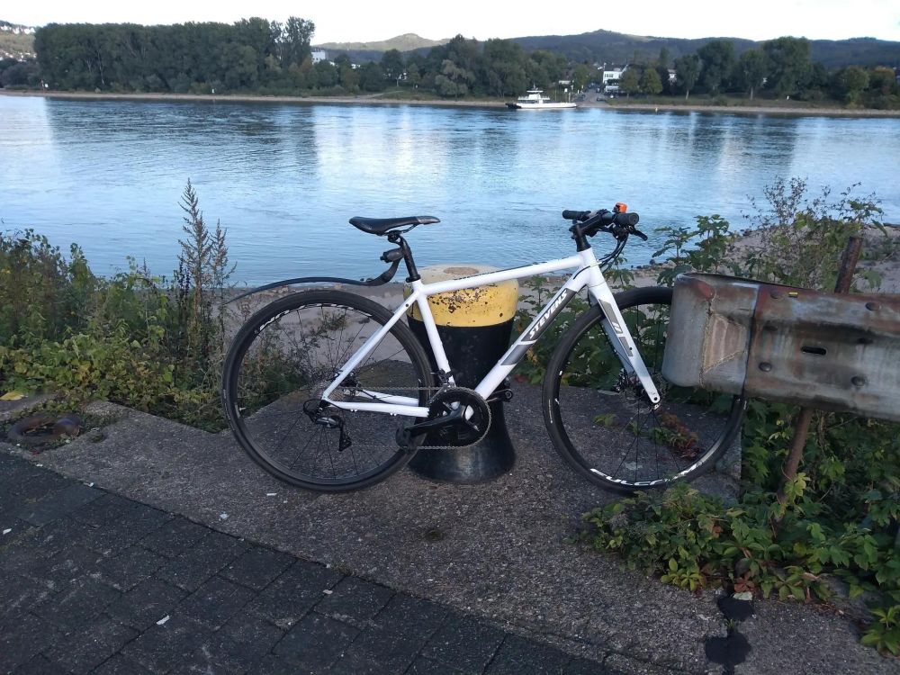 Ein weißer leichter und schneller City-Racer (Fahrrad) vor dem Rhein und im Hintergrund sind Berge zu sehen