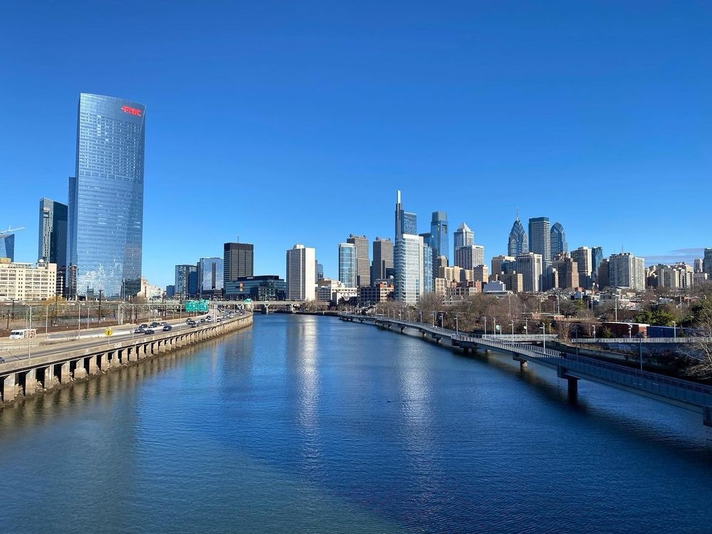 Philadelphia skyline taken from a bridge looking across the river on a sunny day. FMC building is on the left- tall all glass - reflecting blue sky & blue water . Road to the left along the Schuylkill River, shimmering in the middle like a mirror to the sky. Everything dipped in blue: water, buildings, horizon.