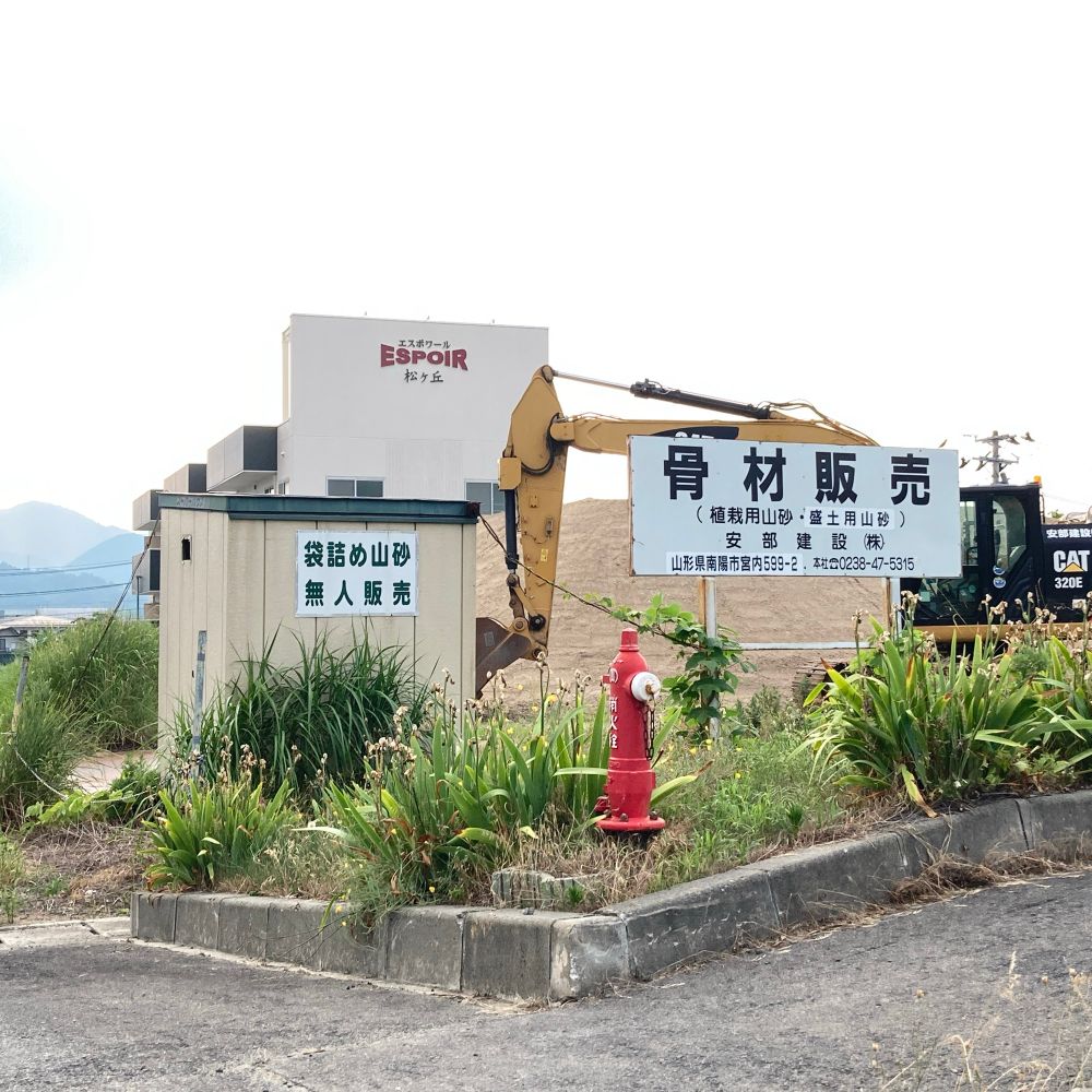 袋詰め山砂無人販売の看板がついた物置小屋と骨材販売の看板後ろにはショベルカー