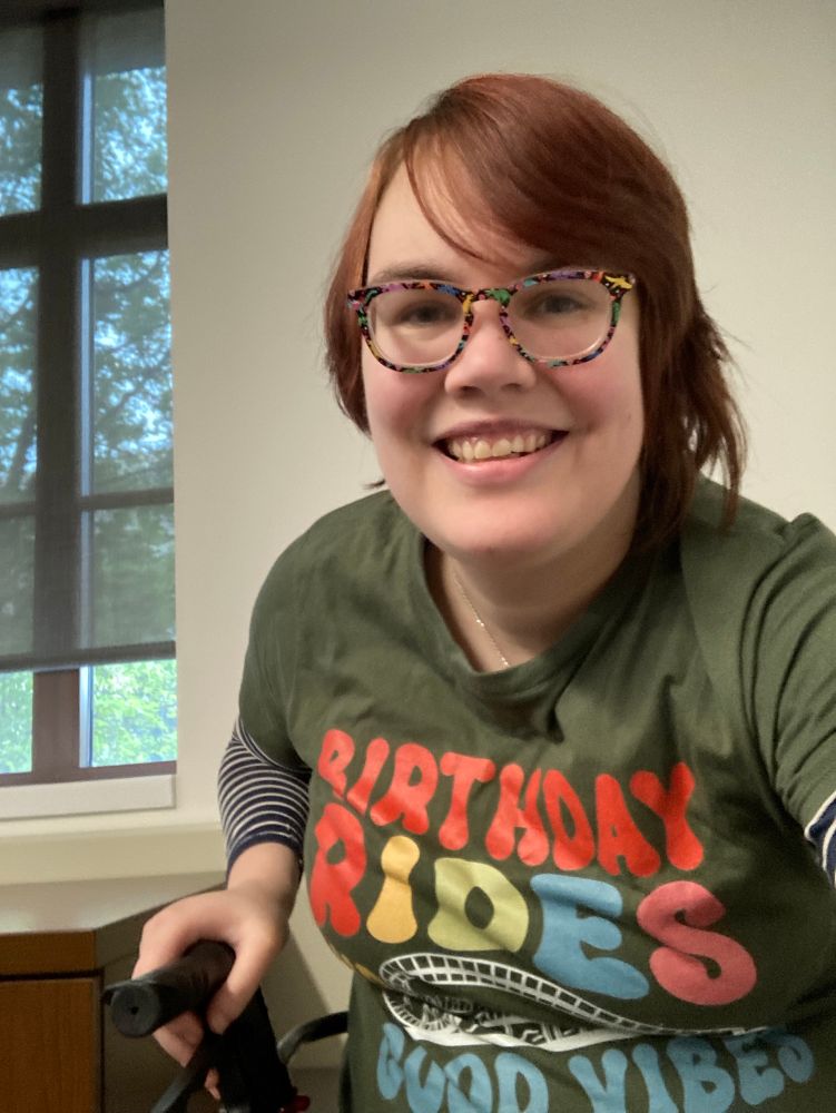 A selfie of a red headed person with glasses frames of dinosaurs with balloons, seated on a rollator with one handle visible in the frame, wearing an olive green t shirt that has a picture of a rollercoaster on it and says BIRTHDAY RIDES AND GOOD VIBES.