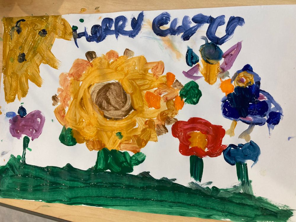 An Easter painting of a sunflower garden with various other flowers, a “cat sun” and a bird my 11 year old daughter painted at the children’s museum, featuring “Happy Easter” in navy cursive. 