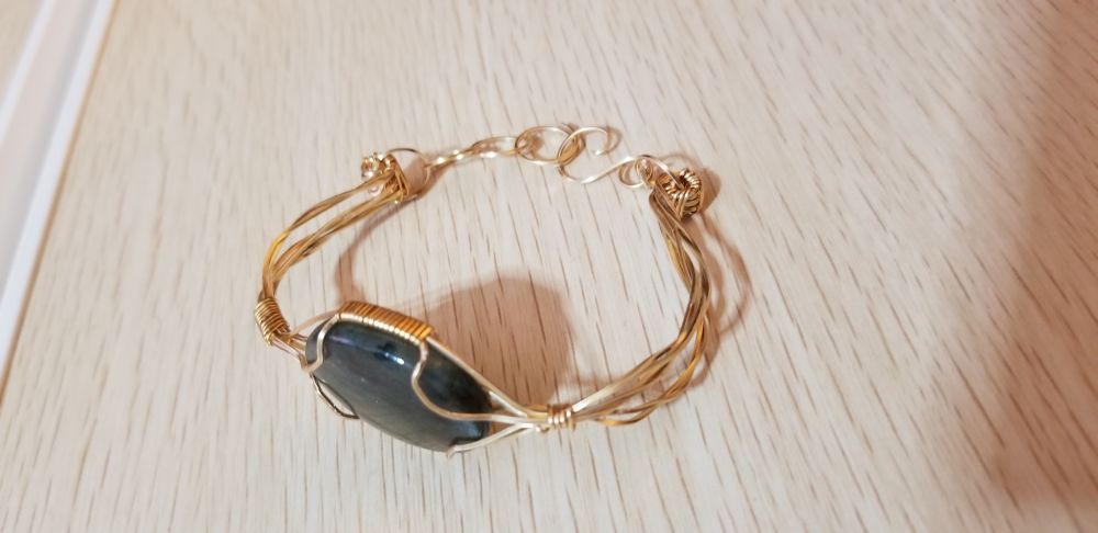 An oval labradorite stone in a gold wire setting. Two twisted wire arms make up part of the cuff, and hand-made jump rings form short chains, with an 'S' shaped hook for a clasp. 