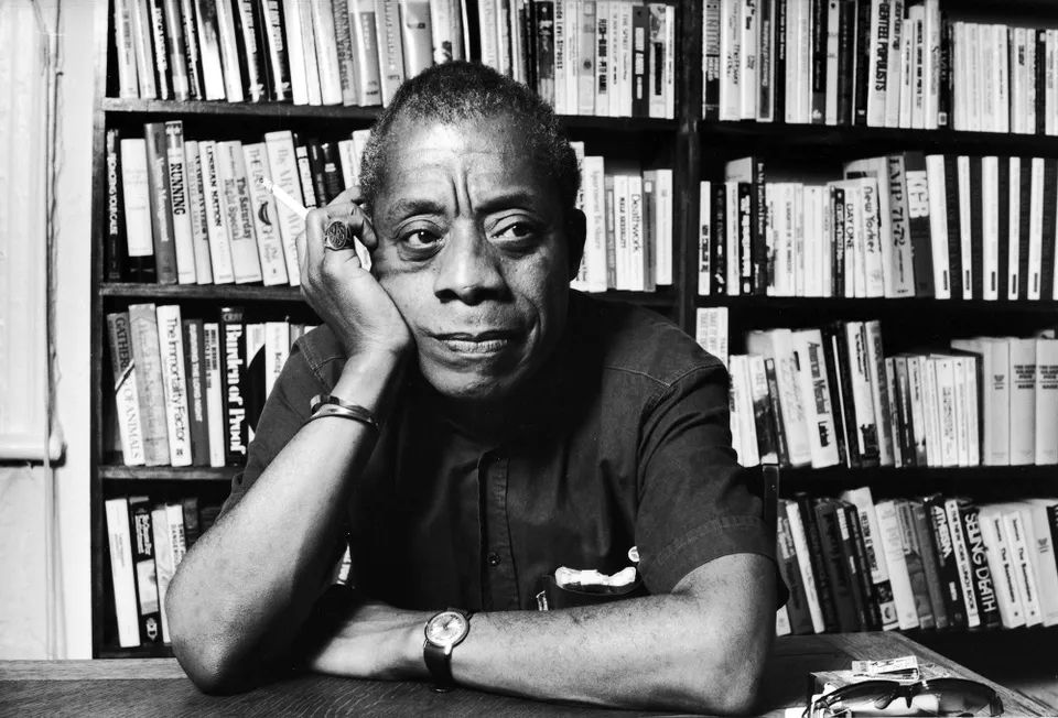 A black-and-white portrait of James Baldwin seated at a table with his head resting on his hand. He is in front of a bookshelf filled with books, wearing a short-sleeve shirt, a watch, and a ring, with a thoughtful expression.