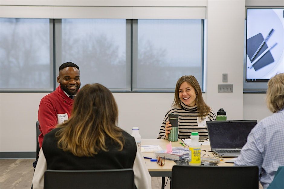 Two people, wearing name tags labeled "Daren" and "Alice," smiling and engaging in a discussion with others around a table in a meeting room with laptops and notepads visible, and a presentation screen in the background.