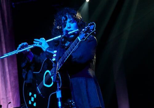 Ann Wilson of Heart playing flute while a guitar hangs from her on a strap. The guitar has a heart shaped sound hole. 
