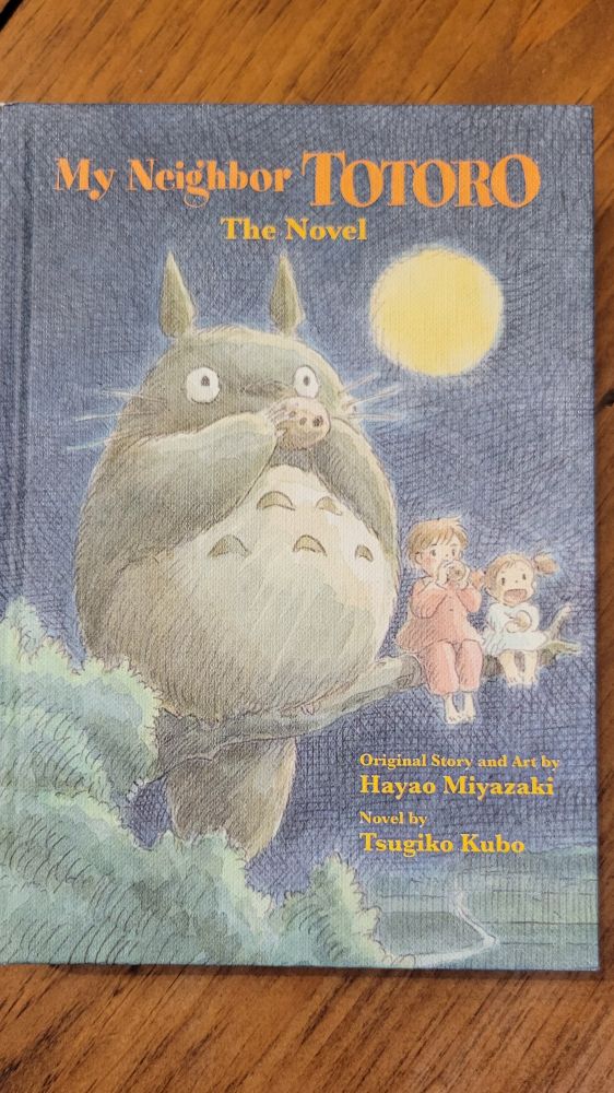 Cover of the novelization of My Neighbor Totoro, featuring artwork of Totoro on a branch with the girls playing flutes.