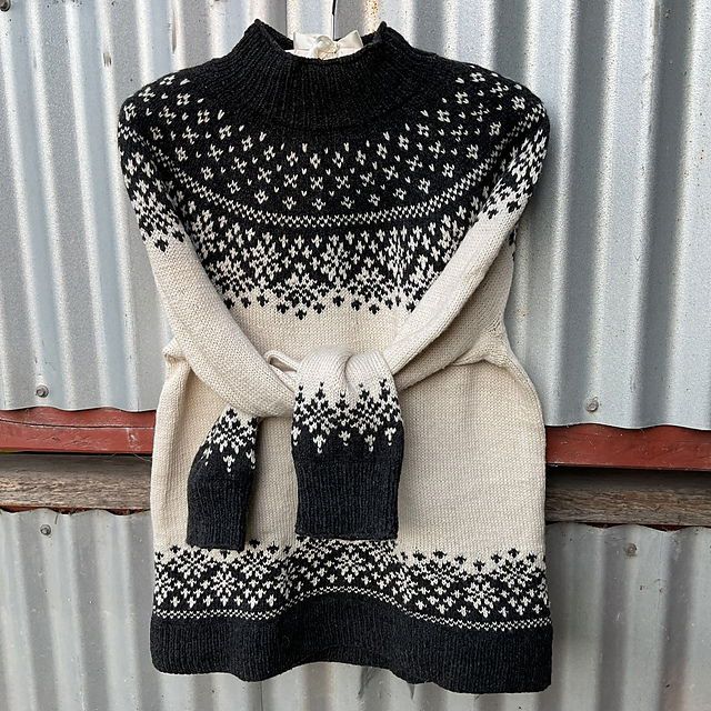 Black and cream sweater with stranded colourwork trim on yoke, lower body and sleeves, hanging against a silver coloured corrugated wall. Sleeves are tied loosely across the body.