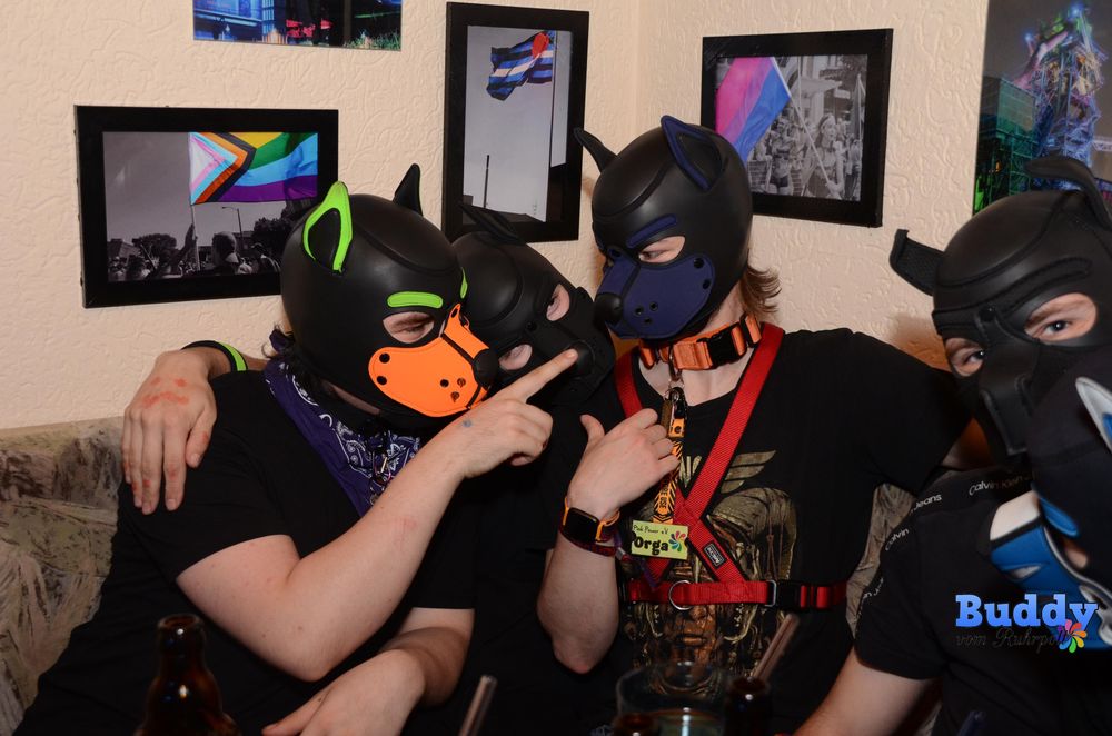 Four people with puppy play masks on sitting on a couch, two of then pointing on a third one and the third one pointing on himself. It looks like the third one is like "What? Me??"