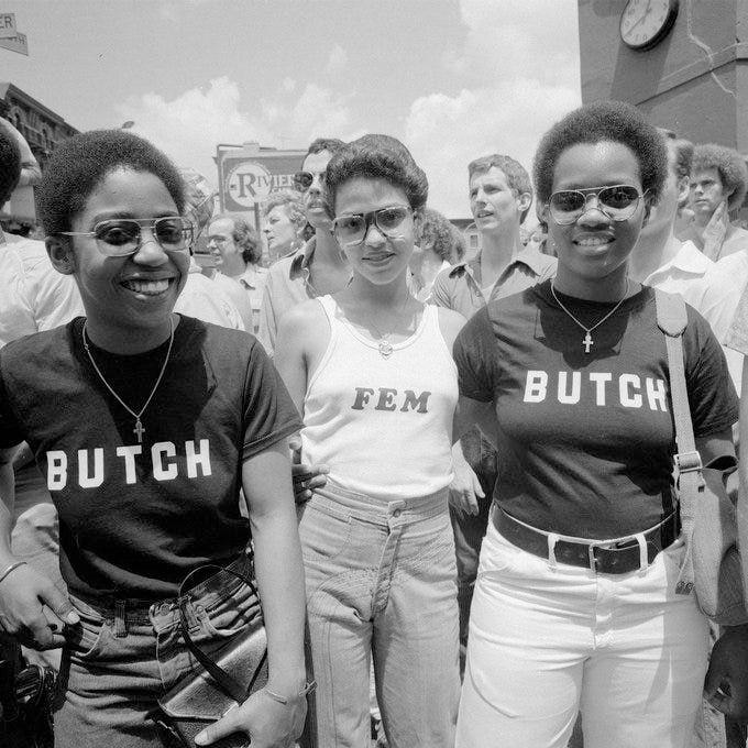 Black and white photo. Three lesbians standing next to each other outside. Two Black lesbians are wearing a black t-shirt that says "BUTCH" on the front. The third lesbian is wearing a white tank top that says "FEM" on the front.