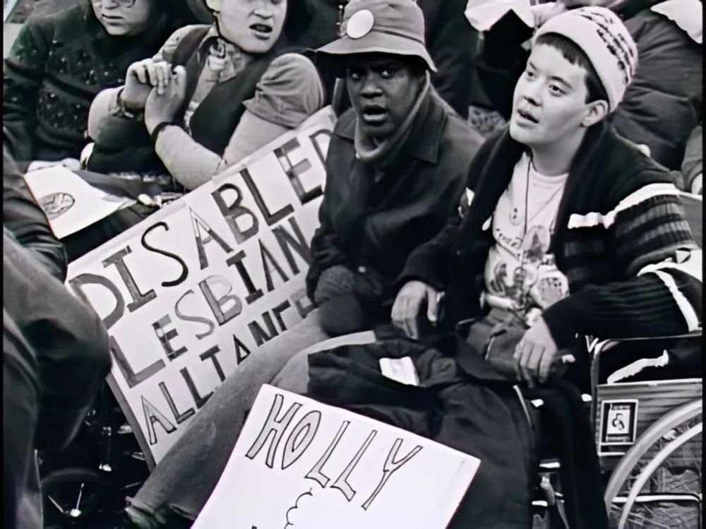Black and white photo. Lesbians in a wheelchair at a protest.  There is a sign in the background that reads DISABLED LESBIANS.