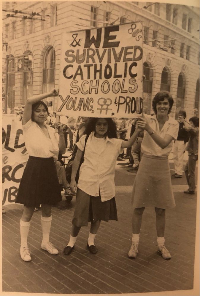 Sepia image of three young femmes dressed as school girls with white blouses and skirts.. They are all holding a sign up that says WE SURVIVED CATHOLIC SCHOOLS. YOUNG, LESBIAN, & PROUD