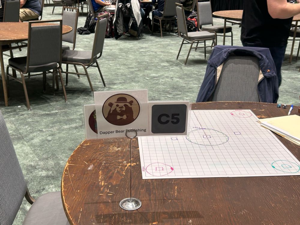 A table standee holds a laminated square with the Dapper Bear logo in it. The logo is a circle with a silhouette of a brown bear wearing a bowler hat and a bowtie.

This photo is somewhat zoomed out & you can see other round tables in the background.