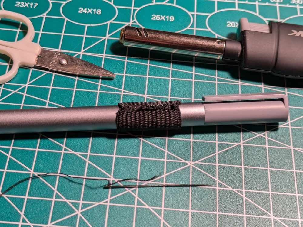 Close-up of the elastic material, fully sewn into a closed loop and test-fitted around my tablet pen.  Also shows the needle and thread, lighter, and some small scissors for cutting the thread.  All on the work mat.