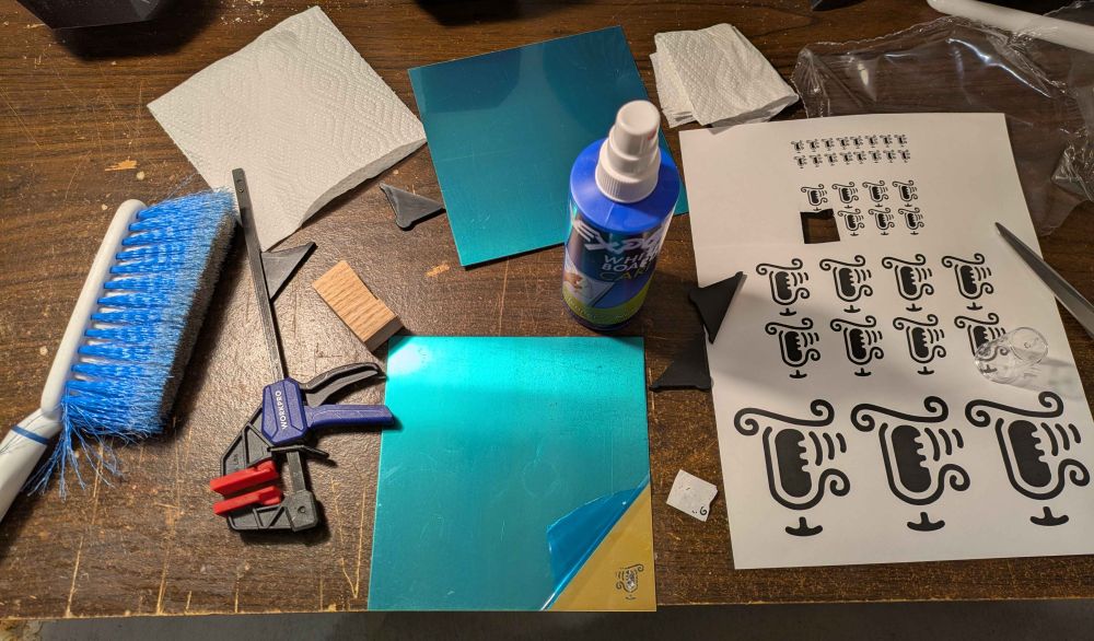 A bunch of mirrored Jerp 'J' logos of different sizes printed on a sheet of paper.  One of the smaller copies is cut out and has already been ~mostly applied to a fresh 6x6-inch sheet of 1mm thick brass.

A small hand clamp, a small block of wood, some paper towels, scissors, and a small dust brush are also visible.