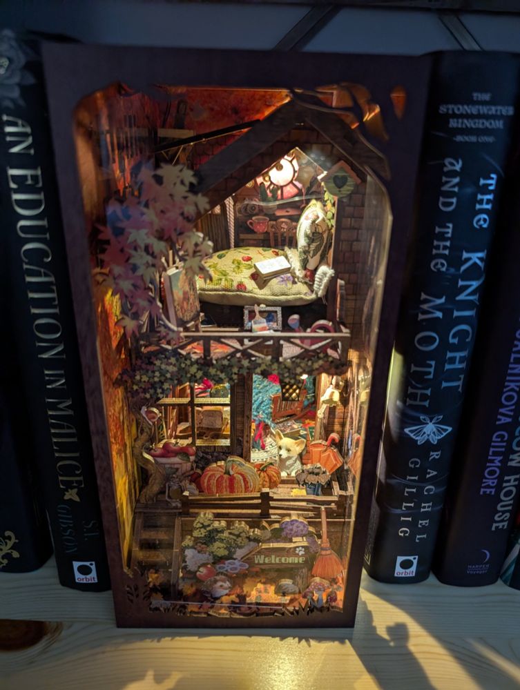 A front view of a tiny house. In the front is a porch with a bunch of pumpkins and plants. A corgi sits in the doorway. Inside is a cosy scene with a loaf of bread just sitting there, and a nice rocking chair with a chunky throw. There's a first-floor balcony with hops and maple leaves, a birdhouse on the front. On the balcony is an easel and paint box. Inside is a bed with an outrageously fluffy duvet and pillow, and a copy of The Invisible Life of Addie Larue waiting to be read. In the far back corner is a desk with a teacup on it. You can't see them but there's other books scattered on the floor. 