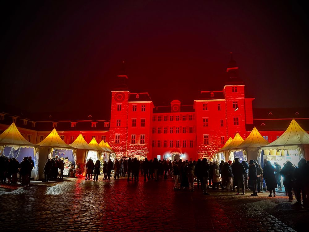 Schwetzinger Schloß bei Nacht, rot beleuchtet. Weinachtsmarktzelte mit Kunsthandwerksstämden im Dunkeln auf dem Platz vor dem Schloß.