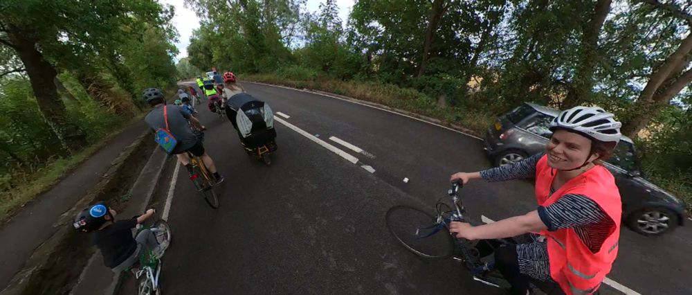 Green county councillor Emma Garnett in pink tabard as a rear marshal in the Abingdon to Europa school bike bus. Ahead are parents and children cycling along the A415 adjacent to the raised causeway footpath. 
