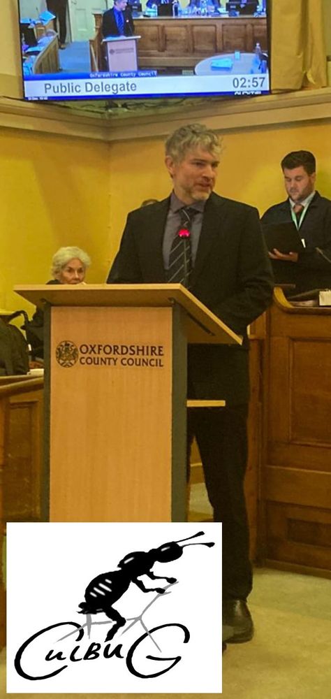Aidan Reilly, uncharacteristically suited, speaking at a lectern at the start of the Oxfordshire County Council meeting. A TV behind shows the 3 minute countdown that public speakers are allowed. A CulBUG logo has been added at bottom left of the photo.