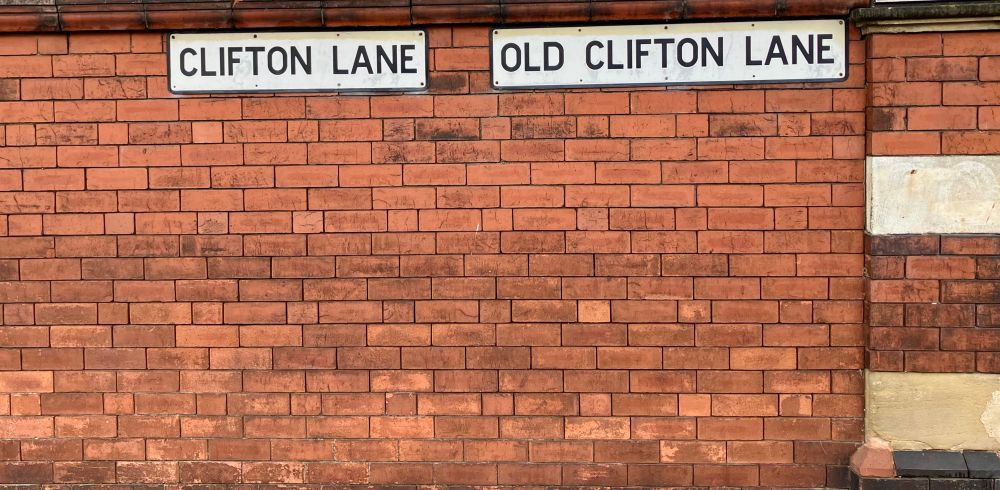Street signs, saying ‘Clifton Lane’ and ‘Old Clifton Lane, on a red-brick wall.