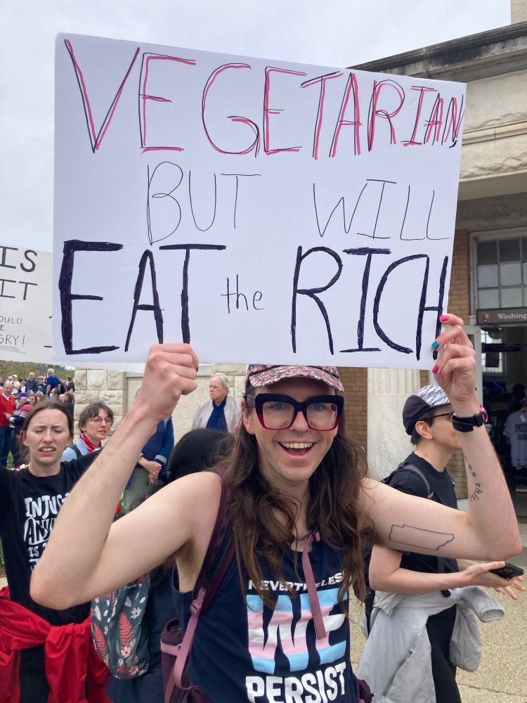 A smiling person holds a sign over their head. It reads “vegetarian but will eat the rich”