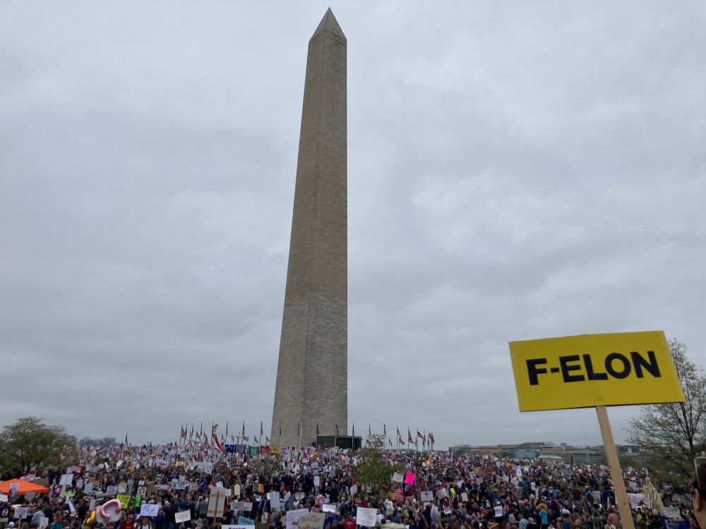 A photo of the Washington monument on a cloudy day. A large crowd is gathered around it. A yellow sign in the foreground reads “F-Elon”