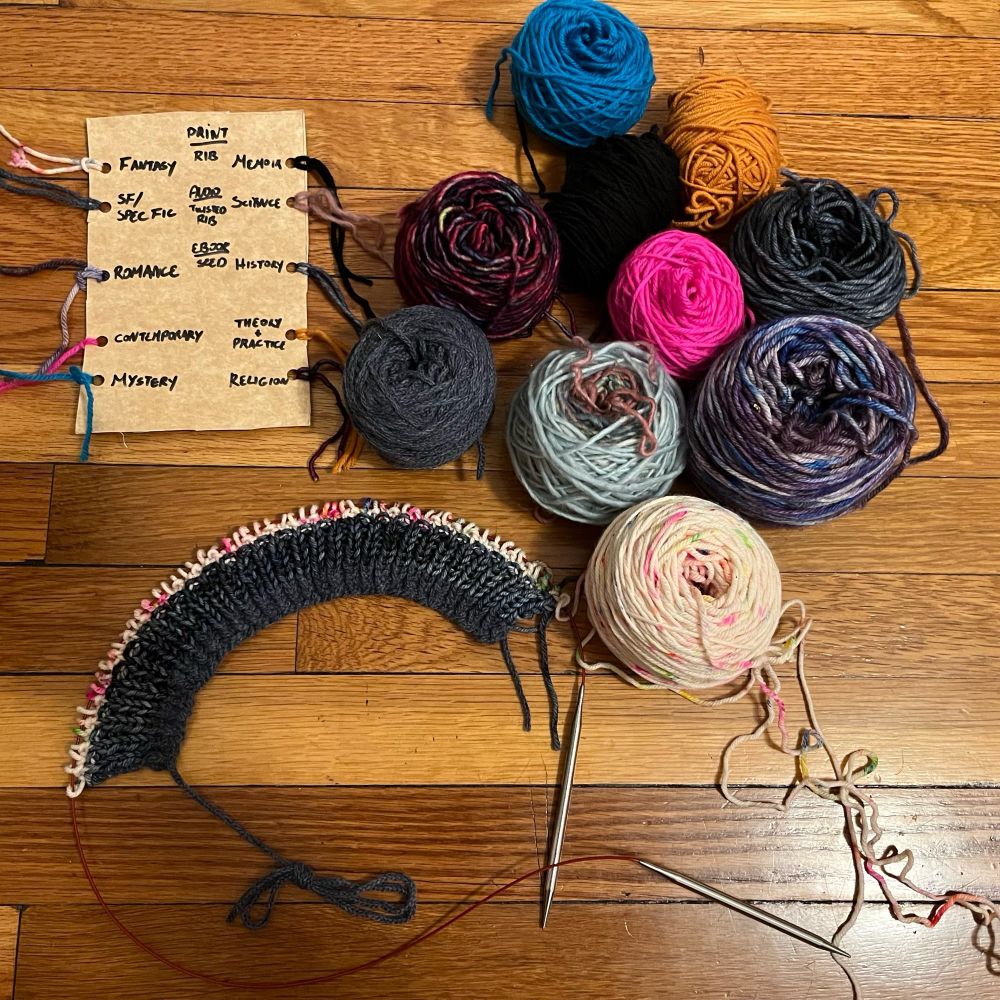 Ten half used skeins of yarn, each a different color, next to a scarf with 12 rows already knit in 3 different colors and patterns. 
There’s also a cardboard swatch card that says which color corresponds to which yarn.