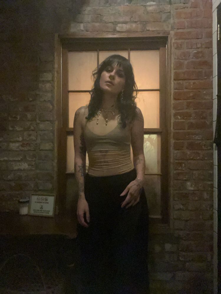 Fera wearing a sheer bodysuit and black slacks, standing against a window frame inside a brick wall, her head tilted to the left looking down at the camera 